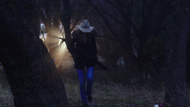 A cowgirl holding a weapon enters the woods quietly, looking for wild animals to hunt, slow motion 23.98 fps.