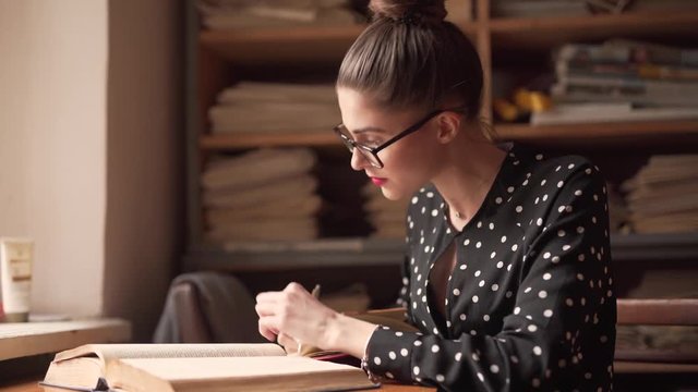 Beautiful librarian leans on pile of books on table in library and sighs. She is miserable and sad as she is not satisfied with ehr job. She raises her eyebrows and creates wrinkles on her forehead.