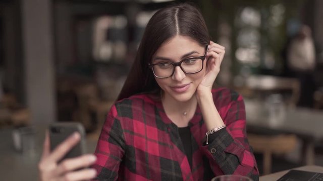 Beautiful brunette girl in red and black checked shirt and glasses sits in restaurant. She takes selfies, smiles, makes kisses, poses and places her hand gracefully under chin. Very blurred background