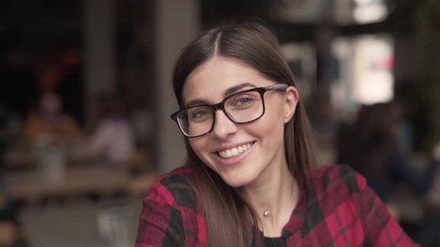 A close up head and shoulder video of beautiful brunette girl in red and black checked shirt. She sits in cafe, smiles from ear to ear and puts on glasses in restaurant. Background is very blurred.