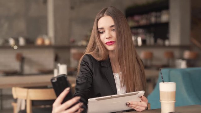 Young Long Brown Hair Lady Girl Sitting in Cafe Holds Tablet in One Hand and Takes Selfie Pictures With Smartphone in Other Hand. Blurred Background