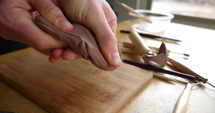Slide across the hands of an artist sculpting with brown modeling clay on a wooden table with tools strewn about the art studio.