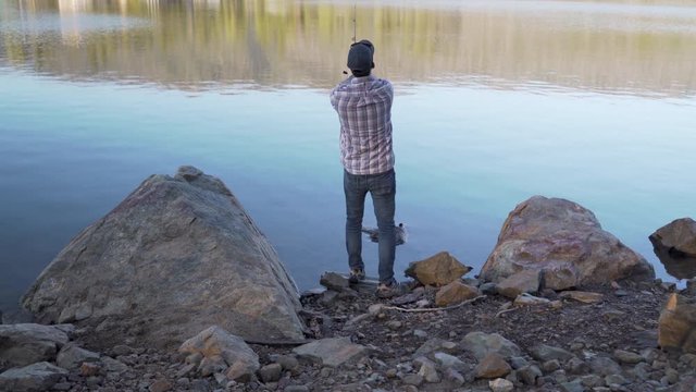 30 Year Old Man Walks Into Left Frame, Casts His Fishing Lure Into A Lake From Rocky Shoreline