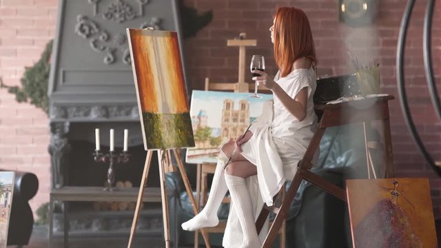 Beautiful artist in a white dress sips from a glass of red wine as she paints in her studio.
