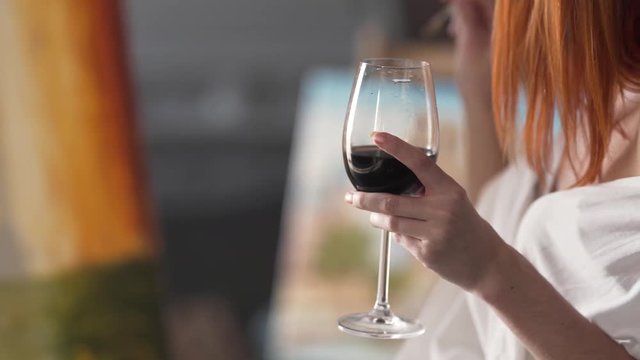A glass of red wine is the focus of the shot, held by a woman who is painting a canvas in her art studio.