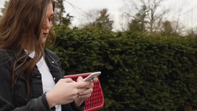 A low angle steadicam shot of a young college student sitting on a bench wearing a white shirt and a black jacket while checking messages on a smart phone.