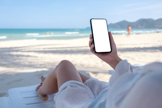 Mockup image of a woman holding white mobile phone with blank desktop screen while laying down on beach chair on the beach