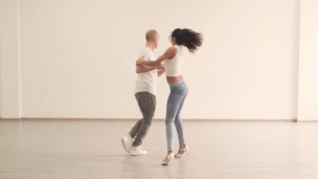 Caucasian Couple Practicing Latin Dance Moves in Studio While He is Circling Aroung Her. They Wearing Light Jeans Pants and White Shirts