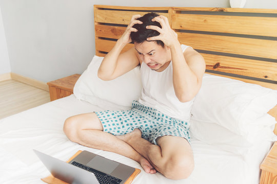 Angry man is working with his laptop on his bed. Concept of freelancer problems.