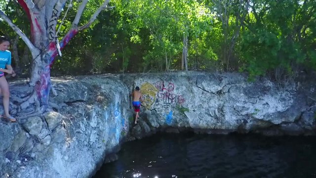 AERIAL: still slow motion shot as a girl prepared to rope swing into the quarry filled water and a boy climbs rocks to escape it.