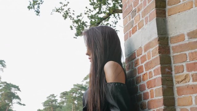 East European brunette mysteriously looking into the camera, surrounded by trees and a brick wall, slow motion footage.