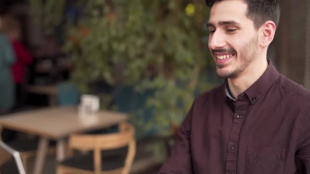 Young Causacian Male Sitting on Terrace of Cafe and Talking To Somebody via Video Call on His Laptop. Blurred Background With People