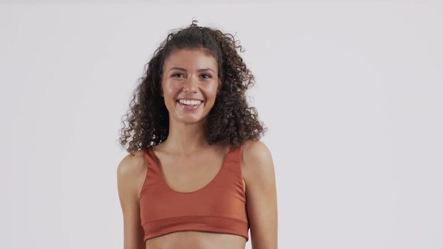 Brunette posing in an orange bikini suit, smiling and playing with her curly brown hair, slow motion footage with a white infinity background.