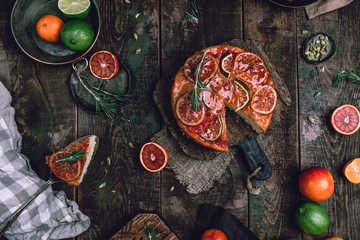 citrus upside down cake with blood oranges, lemons and limes on wooden board among fresh citrus fruit, kitchen stuff, herbs and spices on old wooden background, toned for a vintage effect