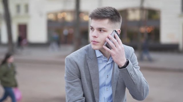 Outdoor Footage of Young Male in Grey Jacket on His Coffe Break Sitting By Street Talking On Smartphone Nodding and Drinking Coffee. Blurred Background and Light Camera Movement