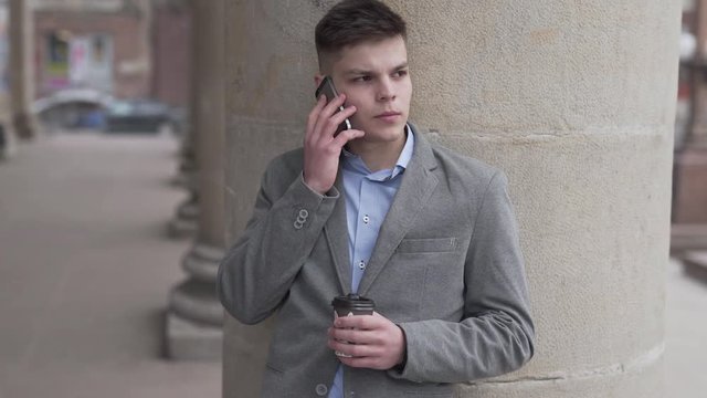 Young Businessman in Grey Jacket and Blue Shirt on His Coffe Break Standing By Building Pole and Talks With Somebody Over Smartphone. Light Camera Movement