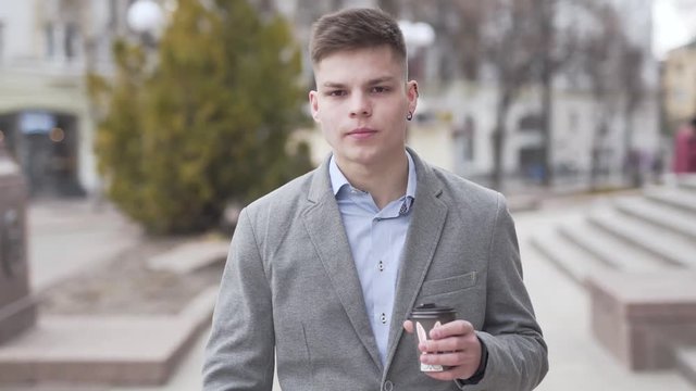 Young Male in Greay Jacket and Earring Walking Down The Street Drinking Coffee To Go and After Checking Smartphone Smiles. Blurred Background
