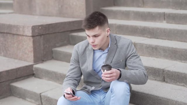 Outdoor Shot of Young Caucasian Male Sitting At Stairs, Drinking Coffee To Go Looking at Smartphone and Then Sky With Smile. Light Camera Movement