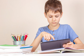Kid sitting at table with books notebooks and using tablet computer