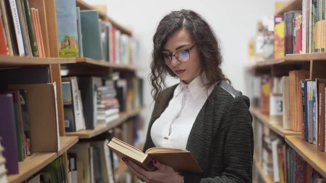 Beautiful young teacher reading a book in a library row with focus, medium shot