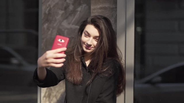 Beautiful young woman stands on the sidewalk in the city using her smartphone to take selfies.