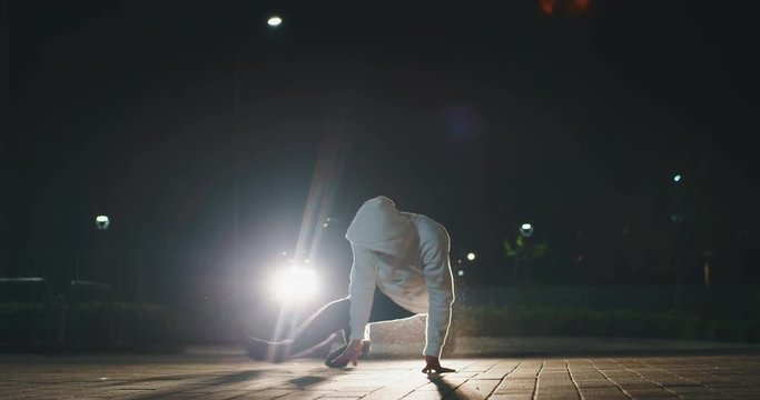 Slow motion of young break dancer is having fun to dancing with car lights at city parking at night.