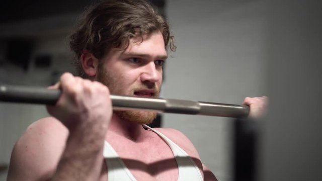 Young bearded ginger male training biceps hard by doing bicep curls with a barbell in an old, rusty Arnold era gym (Gold's Gym) close-up from the front