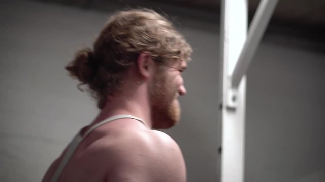 Young bearded ginger male training biceps hard by doing bicep curls with a barbell in an old, rusty Arnold era gym (Gold's Gym) close-up from the back