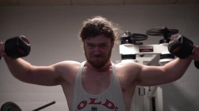 Young bearded male training shoulders hard doing lateral raises with old dumbbells in an old, rusty Arnold era gym (Gold's Gym) close-up