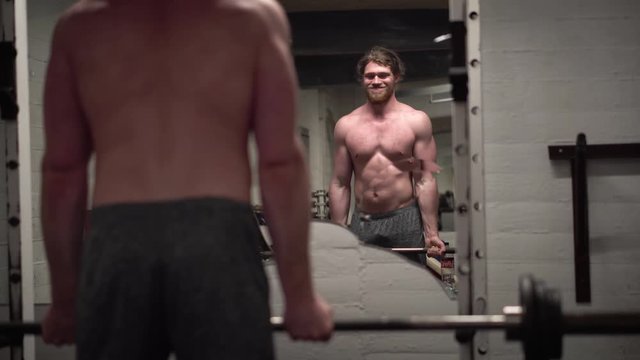 Topless young bearded male showing off and training biceps hard by doing bicep curls with a barbell in front of a broken mirror in an old, rusty Arnold era gym (Gold's Gym) close-up