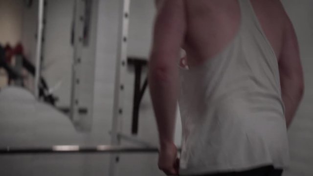 Handheld shaky clip of a young bearded male training shoulders with lateral raises with dumbells in an old, rusty Arnold era gym (Gold's Gym) close-up from the back 4K