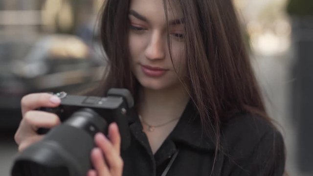 Portrait of Young Caucasian Female in Black Coat and Long Black Hair Holding Photo Camera and Looking At Images on Display With Smile. Blurred Background Light Camera Movement