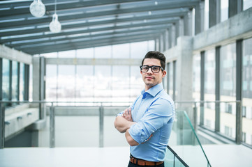 A portrait of young businessman standing indoors in an office, arms crossed.