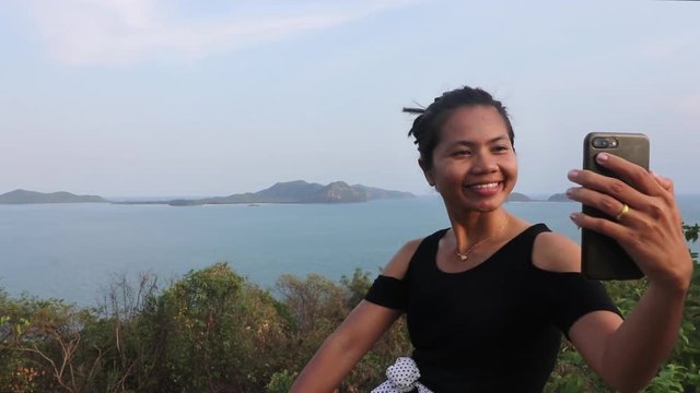 A steady close-up shot of a young thai girl who is taking herself selfy photos at a beautiful viewpoint with islands and the andaman sea behind her, Thailand.