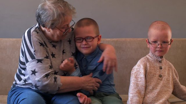 elderly woman sits and plays with her grandchildren on couch. Children love grandmother very much. Boys kiss and hug their beloved grandmother
