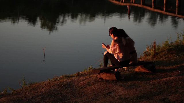 Angled view of two girls sitting on a bench by peaceful lake with reflections from trees in the golden hour and checking phone