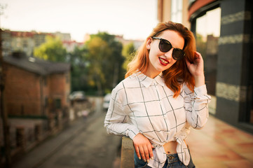 Attractive redhaired woman in sunglasses, wear on white blouse posing at street against modern building.