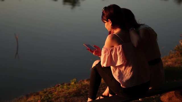 Angled backside view of two girls sitting on a bench by peaceful lake in the golden hour