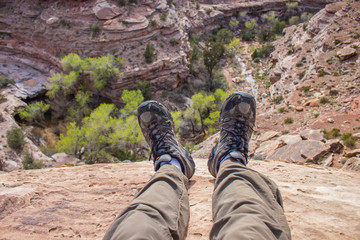 A hiker dangles his feet over the edge of a cliff looking down into a desert canyon in this first person pov photo.  - obrazy, fototapety, plakaty