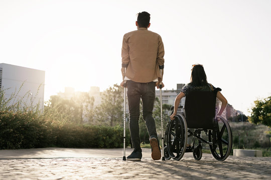 Back view of woman on wheelchair and her boyfriend using crutches