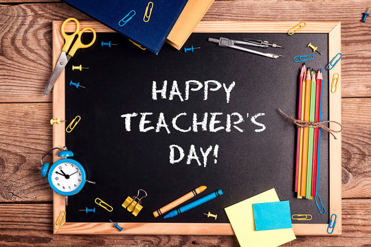 A chalkboard with the text happy teachers day written in it, school supplies and alarm clock on a rustic wooden table.