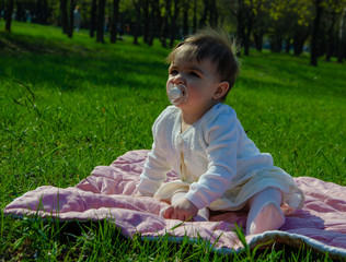 Baby in bright clothes on a pink plaid on green grass in the park. Newborn baby outside on a warm day. Little girl plays on a plaid on a green lawn in spring