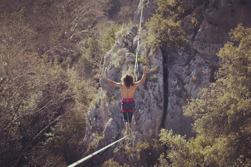 Obraz premium A man is walking along a stretched sling. Highline in the mountains. Man catches balance. Performance of a tightrope walker in nature. Highliner on the background of valley.