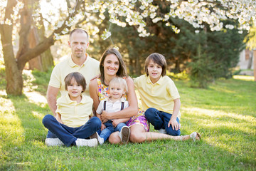 Fototapeta premium Piękna rodzina, matka, ojciec i troje dzieci, chłopców, o rodzinnym portret na zewnątrz zrobiony w słoneczny wiosenny wieczór, piękny kwitnący ogród, zachód słońca