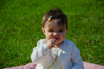 Fototapeta premium Baby in bright clothes on a pink plaid on green grass in the park. Newborn baby outside on a warm day. Little girl plays on a plaid on a green lawn in spring