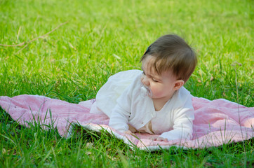 Naklejka na ściany i meble Baby in bright clothes on a pink plaid on green grass in the park. Newborn baby outside on a warm day. Little girl plays on a plaid on a green lawn in spring