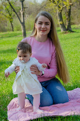 Fototapeta premium Mom with baby in bright clothes on a pink plaid on the green right. Family resting in the park on a warm day. Mom and little girl 10 months walk in the park
