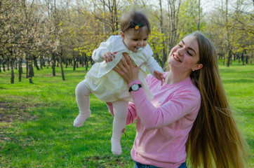 Mom with baby in bright clothes on a pink plaid on the green right. Family resting in the park on a warm day. Mom and little girl 10 months walk in the park