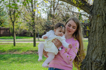 Mom with baby in bright clothes on a pink plaid on the green right. Family resting in the park on a warm day. Mom and little girl 10 months walk in the park