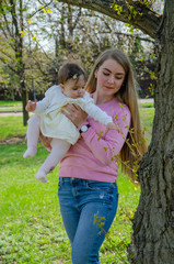 Mom with baby in bright clothes on a pink plaid on the green right. Family resting in the park on a warm day. Mom and little girl 10 months walk in the park